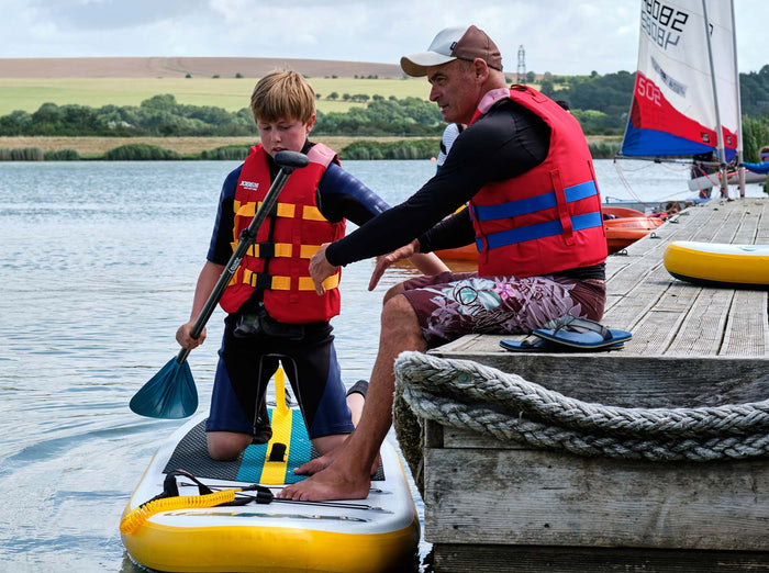 Watersports instructor helping child get on SUP board on lake